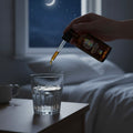 Hand pouring liquid from a dropper bottle into a glass on a bedside table at night.