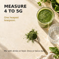 A heaped teaspoon containing 4 to 5g of green powder beside water and a green smoothie, illustrating suggested use.