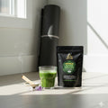 Glass of green juice and a package of Ormus Gold Greens on a light surface with a white wall and window in the background.