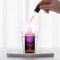 Hand using a dropper to add liquid from a purple bottle into a glass on a white background