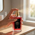 Hand holding a red and gold bottle of 'Gold Gold' face and body mist on a wooden surface.