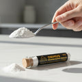 Hand holding a spoon with white powder above a container labeled 'Ormus Monatomic Gold' on a light surface.