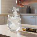 Glass of water with dry ice and a spoon, with ORMUS Monatomic Gold product on a kitchen counter.