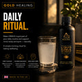 Gold Healing Liquid Ormus Monatomic Gold daily ritual supplement bottle and a glass of cloudy liquid on a dark tabletop with warm background lighting.